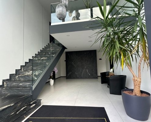 Dieses Bild zeigt ein modernes Entree. Der Boden ist mit großen hellen Fliesen gefliest. Im hinteren Teil befindet sich eine graue Wand in Mamor. Darüber befindet sich eine Empore die über eine Treppe aus grauen Mamor zu erreichen ist. Im voderen Teil findet sich ein Teppich mit der Aufschrift Gemeinsam Zukunft gestalten. Am rechten Bildrand steht eine Palme und dahinter eine Sitzgruppe.