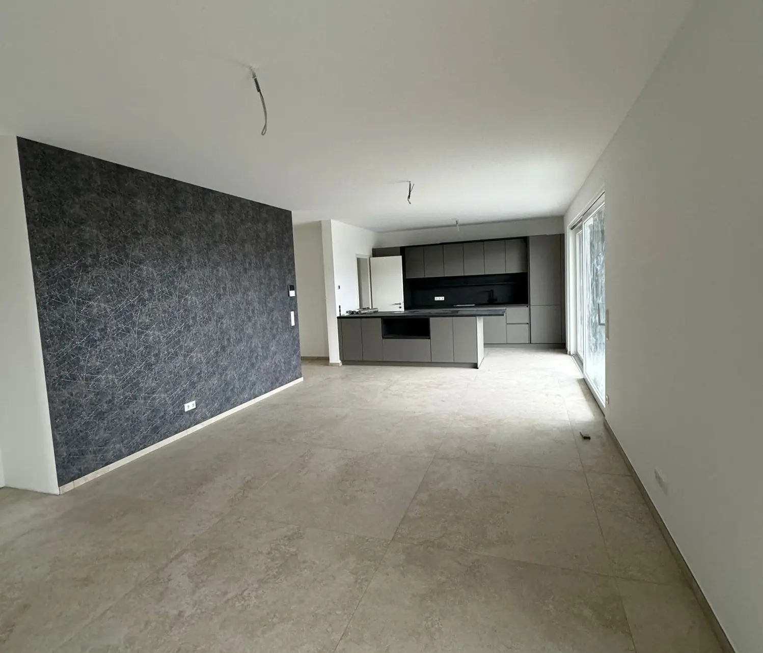 Raum mit Kueche Objekt Aachen Zu sehe ist hier ein fertig renovierter Raum. Die linke Bildseite zeigt eine grau gemusterte Wand. Am ende des Raumes ist eine moderne grau Küche mit Kochinsel zu sehen.